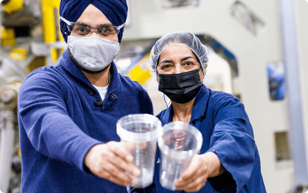 2 smiling workers in factory holding up clear drink containers to camera