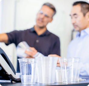 foreground clear drinking cups of different sizes, background blurred image of 2 office workers talking