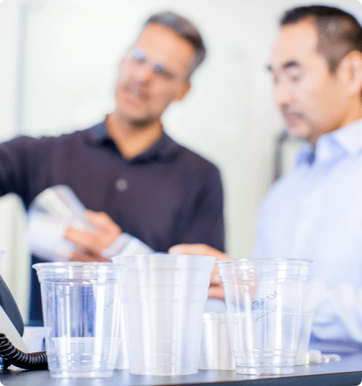 foreground clear drinking cups of different sizes, background blurred image of 2 office workers talking